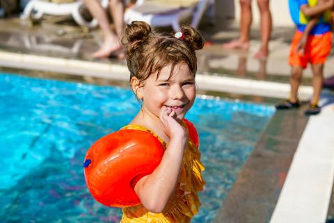toddler in bright orange yellow swimming costume and armbands on, stood by a swimming pool