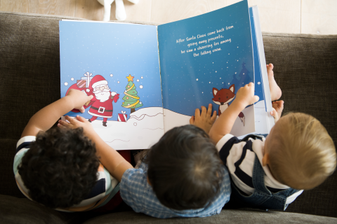 Birds eye view of thee children reading a book - the book is blue and has an image of Santa Claus on the left hand side. You can see the tops of the three children's heads, and their hands holding the book together.