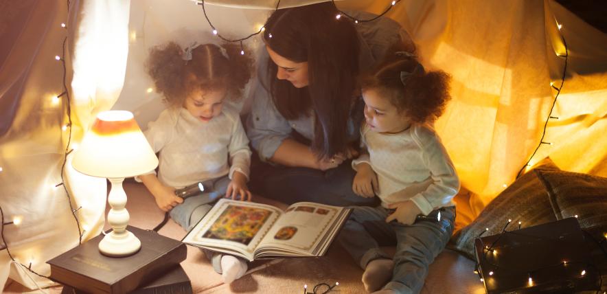 Reading and family games in children's tent. Mother and two twins daughters with books and flashlight before going to bed.