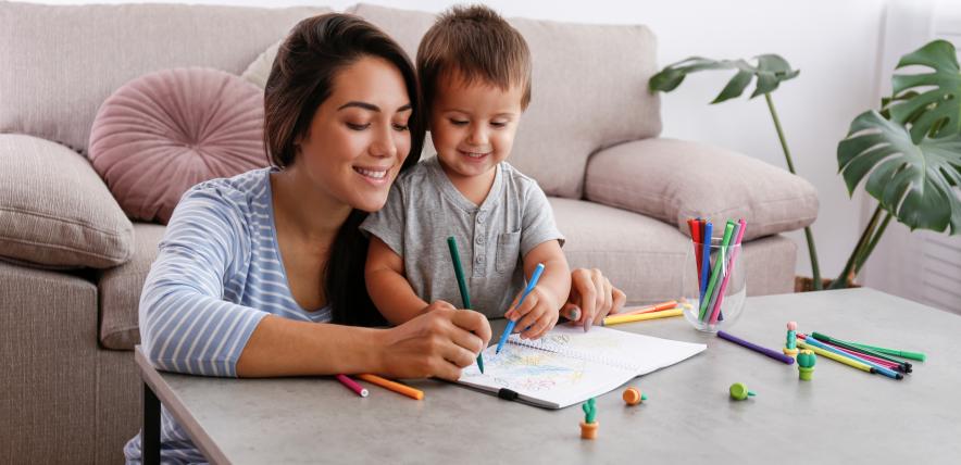 Mother and young boy sit on the floor at a table, drawing with coloured pencils and paper