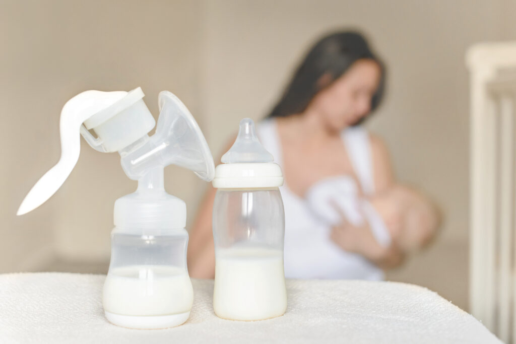 Close-up of breast bump and bottle half-filled with milk, with mother breastfeed her baby out of focus in the background