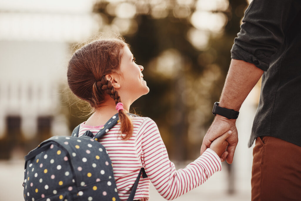 Young girl with backpack looking up at her father smiling and holding his hand