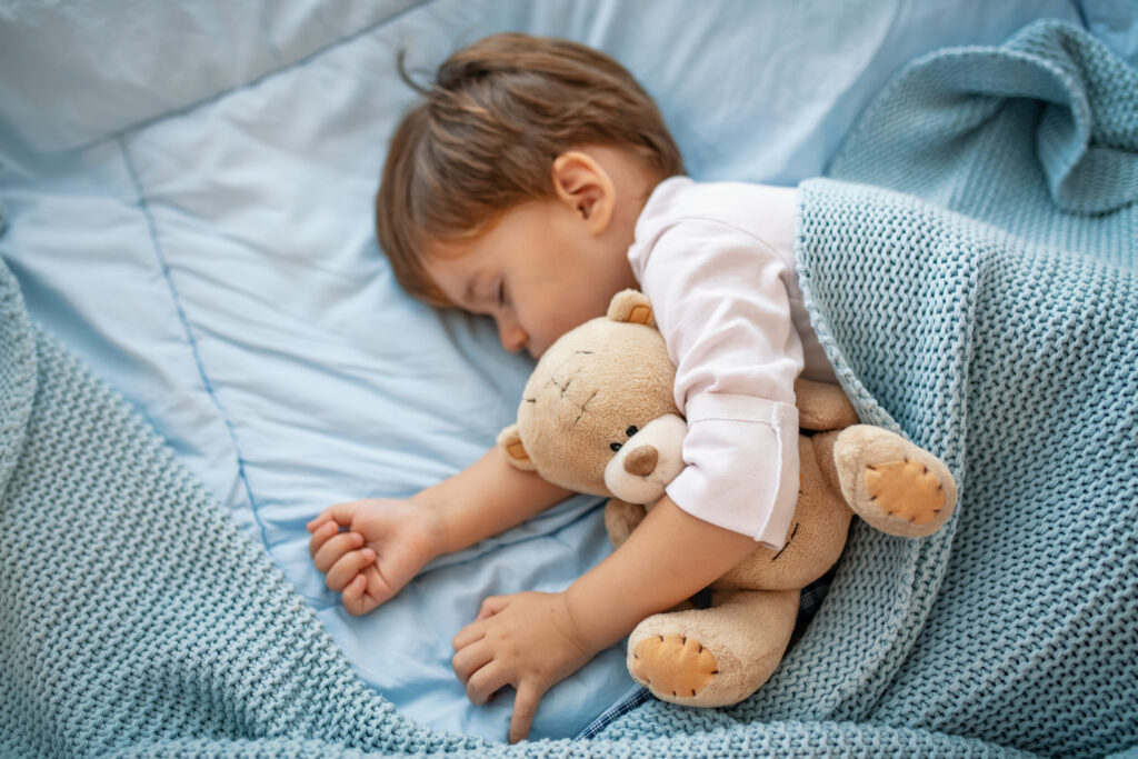 Child tucked into beed with blue sheets and a blanket, and a teddy bear tucked under their arm