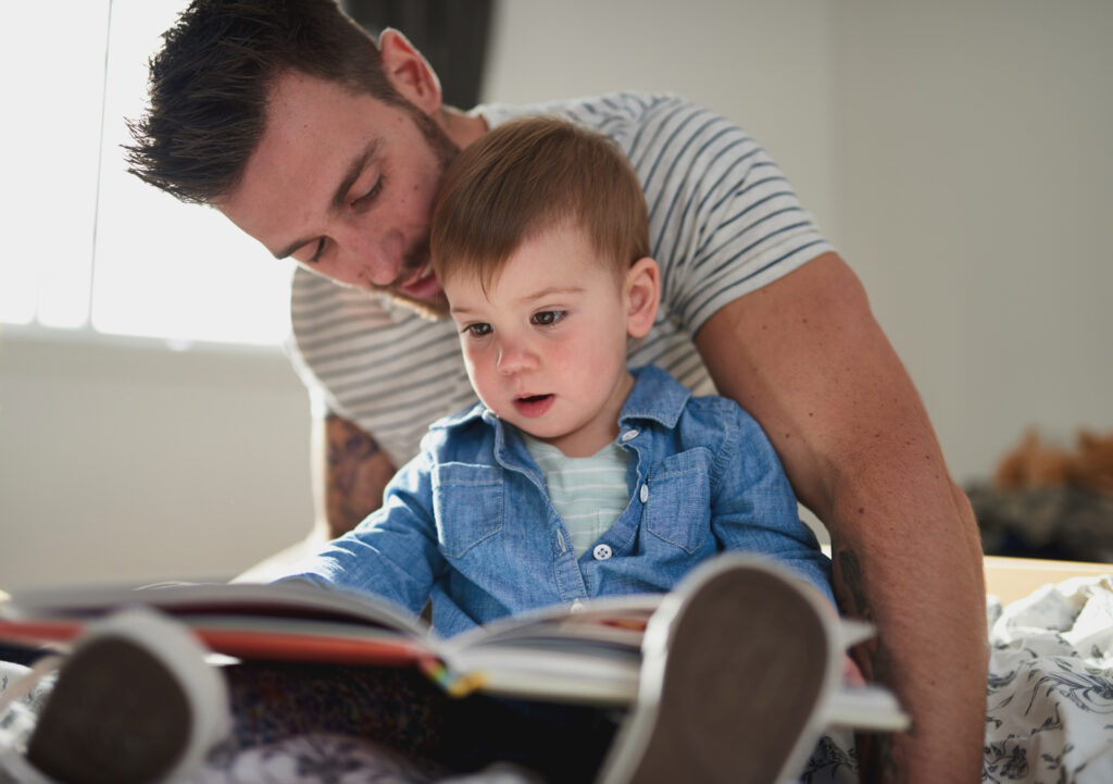 A man read a book with a young boy - the boy wears a button down blue shirt, and the man wears a striped tshirt