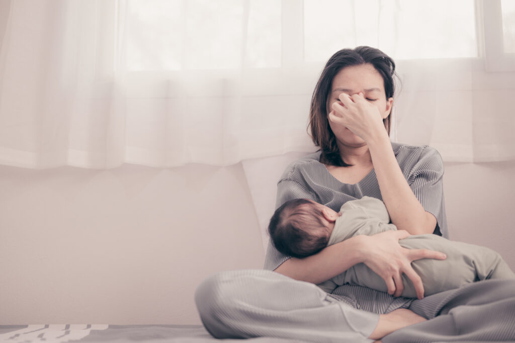 Stressed mother pinching her nose sat by the window on the floor with a small baby in her arms