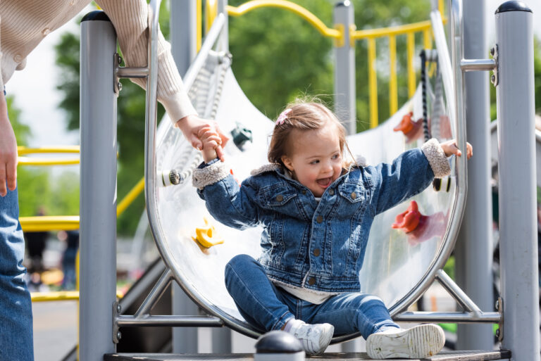 Toddler in denim jacket and jeans holds an adults hand who is out of the shot while getting off the bottom of a metal slide