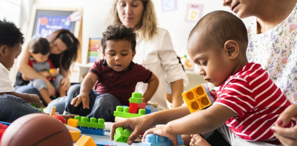 Two toddlers in the foreground play with coloured building bricks, sitting on the laps of two women. Another two children and one woman are visible in the background of the image, also playing with toys.